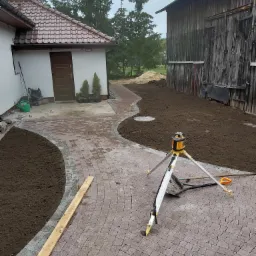 Świeżo ułożona, łukowata ścieżka z kostki brukowej w odcieniach czerwieni i szarości, obok przygotowany teren pod trawnik, widoczny niwelator laserowy na statywie i narzędzia brukarskie, w tle...