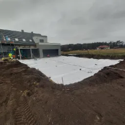 Przygotowanie terenu pod budowę tarasu: wykop z białym materiałem izolacyjnym, widoczny budynek w trakcie wykończenia i pracownicy w odblaskowych kamizelkach.