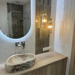 GTO GROUP Andrzej Liczbiński - Nowoczesna łazienka z kamienną umywalką, podświetlanym lustrem i wiszącymi lampami. Brązowe płytki w strefie prysznica i nad blatem. Drewniana szafka pod umywalką.