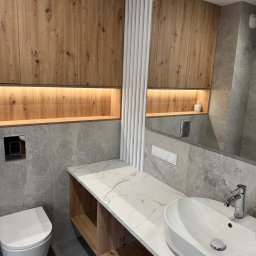 GTO GROUP Andrzej Liczbiński - Nowoczesna łazienka z drewnianymi szafkami, marmurowym blatem i umywalką nablatową. Szare płytki i białe lamele dekoracyjne.