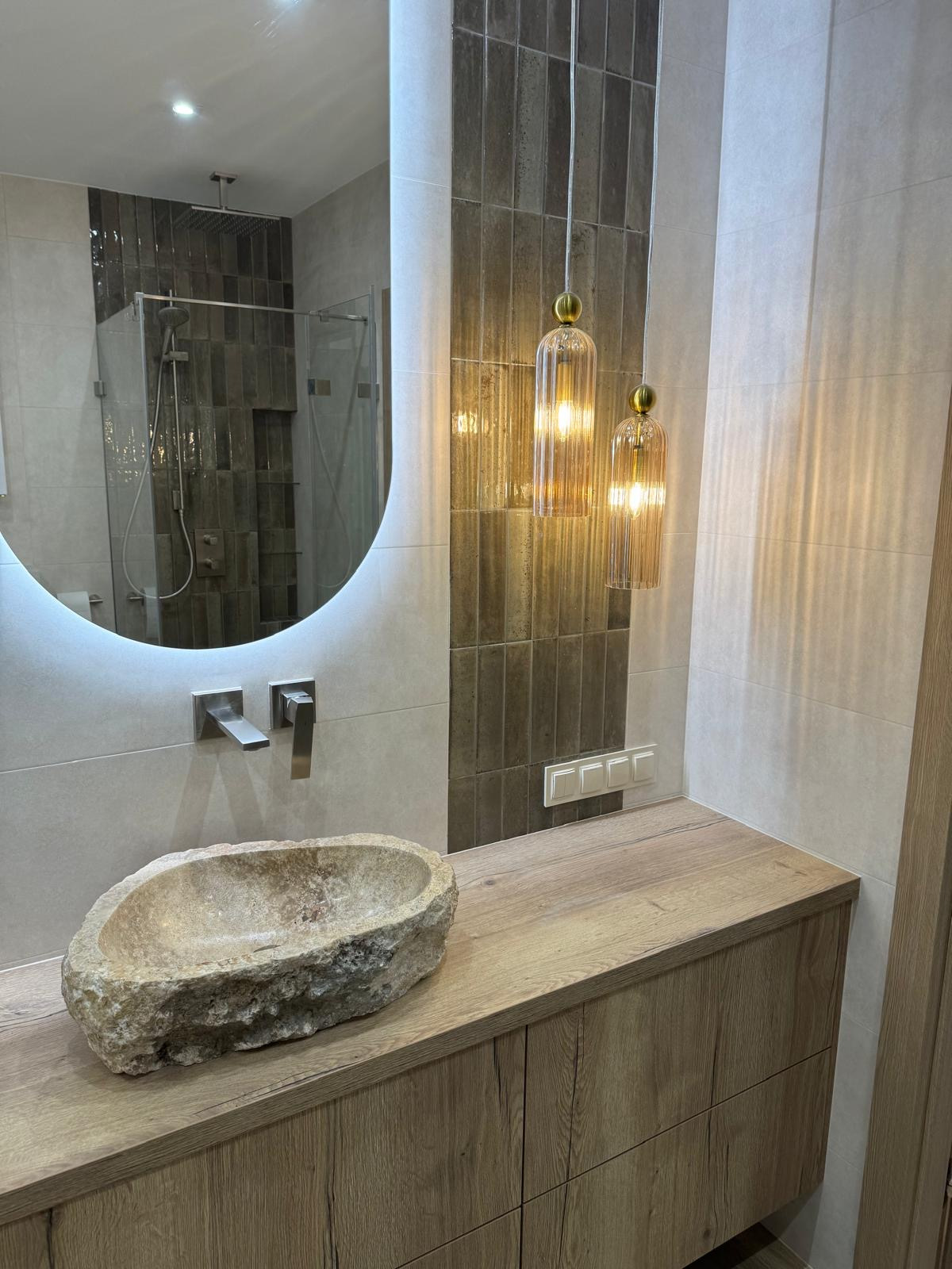 Nowoczesna łazienka z kamienną umywalką, podświetlanym lustrem i wiszącymi lampami. Brązowe płytki w strefie prysznica i nad blatem. Drewniana szafka pod umywalką.