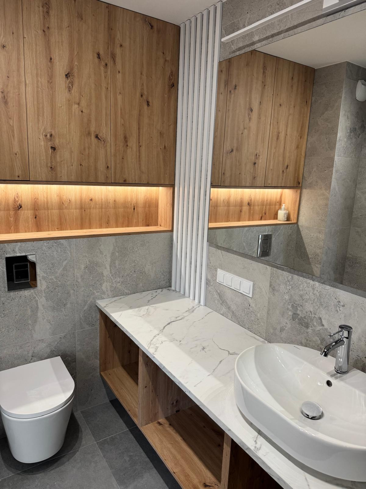 Nowoczesna łazienka z drewnianymi szafkami, marmurowym blatem i umywalką nablatową. Szare płytki i białe lamele dekoracyjne.