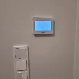 Panel sterowania alarmem Satel zamontowany na ścianie obok włączników światła, wyświetlający ikony kłódek otwartej i zamkniętej.