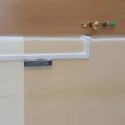 Klawiatura numeryczna domofonu z szarym panelem, zamontowana na kremowej ścianie obok tabliczki z napisem 'Archiwum 06', widoczne pęknięcia na ścianie.