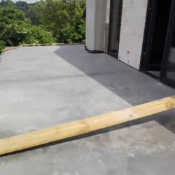 Szara, gładka powierzchnia posadzki na tarasie budynku w trakcie prac wykończeniowych. W tle widoczna ściana z bloczków i zieleń drzew.