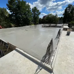 Świeżo wylana, gładka, szara płyta betonowa z wystającymi prętami zbrojeniowymi, widok z góry, w tle zielone drzewa i błękitne niebo z chmurami.