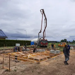 Budowa fundamentów domu jednorodzinnego: szalunek z drewna, beton pompowany z betoniarki, pracownicy w trakcie układania bloczków betonowych, pochmurne niebo.