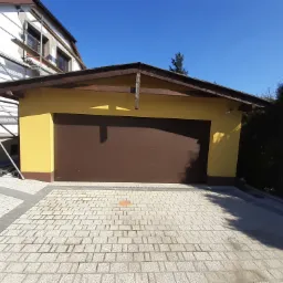 Żółty garaż z brązową bramą segmentową, widoczny fragment domu w remoncie ze rusztowaniem, kostka brukowa na podjeździe.