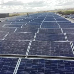 Rząd paneli słonecznych na dachu budynku przemysłowego, widok z góry, pochmurne niebo w tle.