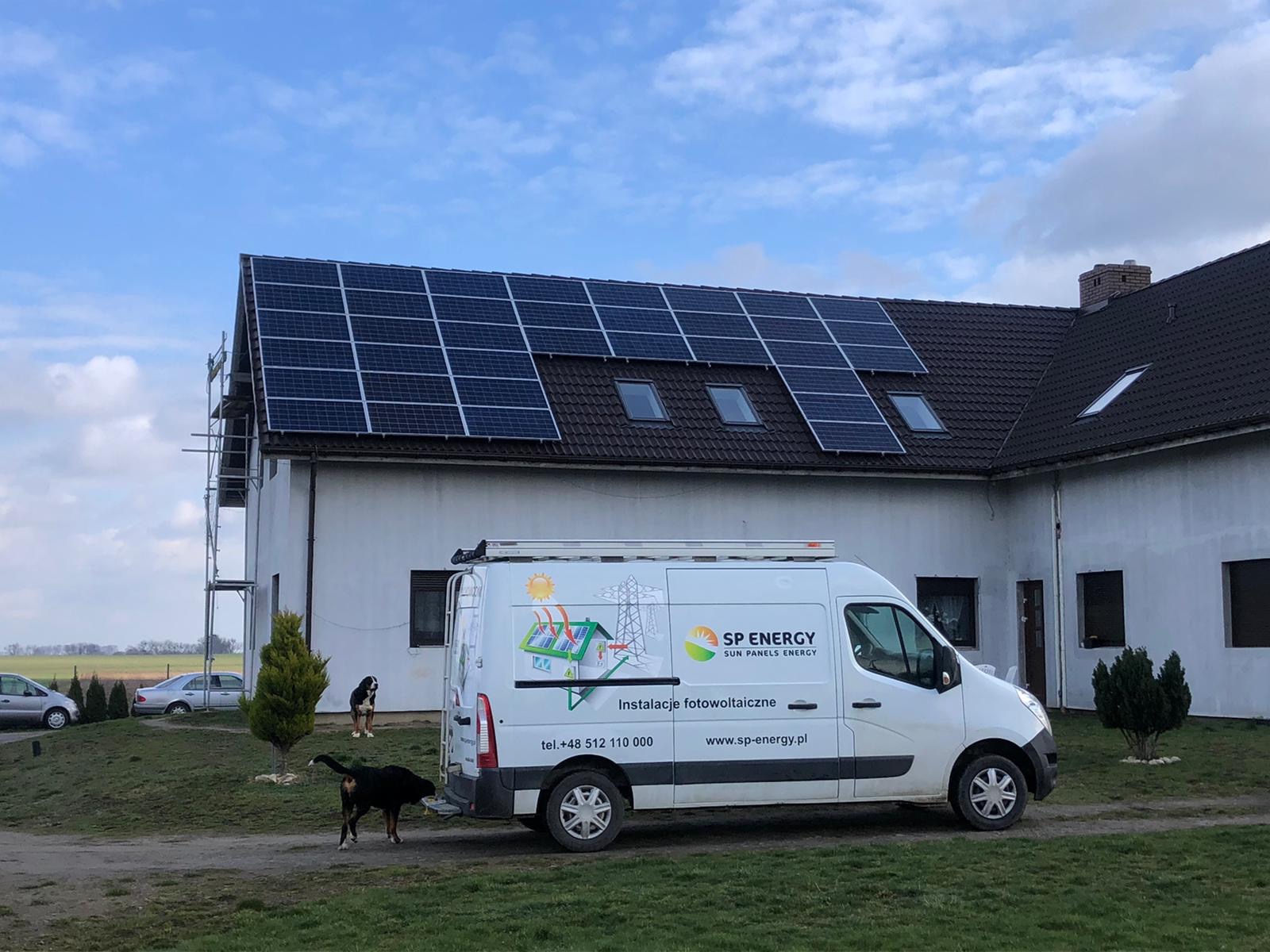 Dom z panelami słonecznymi na dachu, częściowo zamontowanymi, widoczne rusztowanie, na podwórku biały bus firmy SP Energy oraz dwa psy.