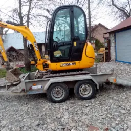 Żółta minikoparka JCB 8018 CTS umieszczona na szarej przyczepie, stojąca na żwirowym podłożu, z budynkami i drzewami w tle.