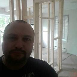 Vasyl - Ujęcie z dołu na drewnianą konstrukcję ścianki działowej w trakcie budowy w jasnym pomieszczeniu, widoczny fragment twarzy wykonawcy.