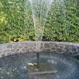 Vasyl - Okrągła fontanna z kamienną obudową i zielonym żywopłotem w tle, woda tryskająca z dyszy w górę.