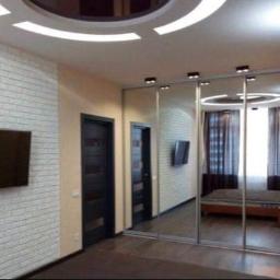 Vasyl - Nowoczesne wnętrze pokoju z białą ceglaną ścianą, telewizorem, przesuwną szafą z lustrami odbijającymi łóżko i okno, oraz designerskim oświetleniem sufitowym.