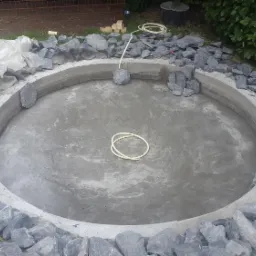 Betonowy okrągły zbiornik w trakcie budowy, obłożony szarymi kamieniami, widoczne narzędzia i materiały budowlane w tle.