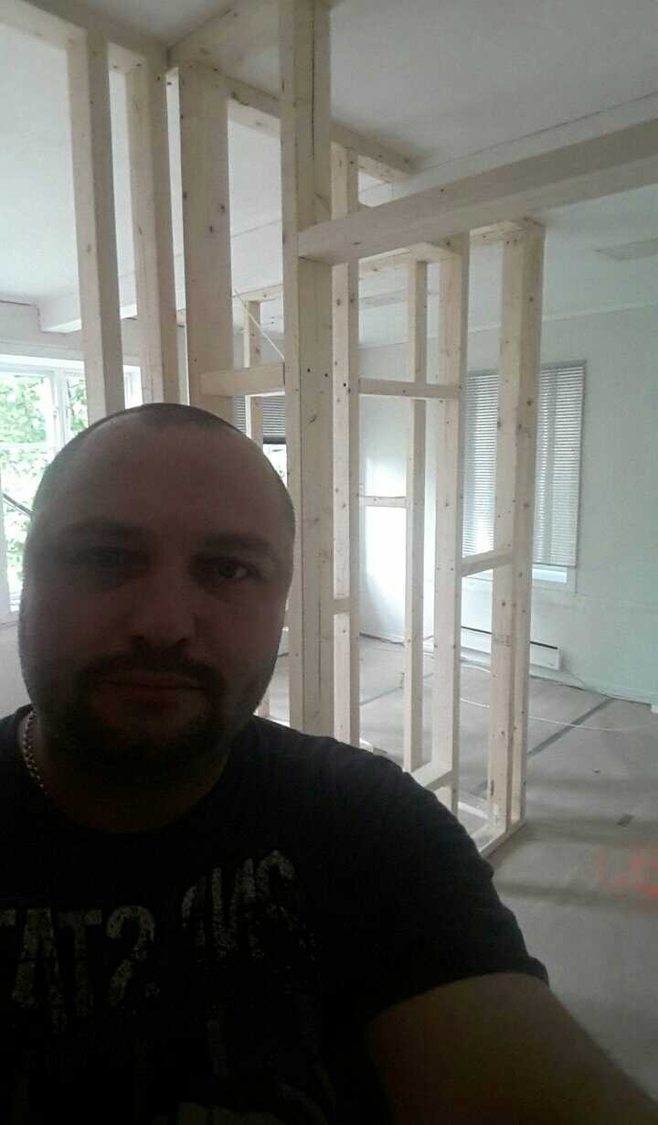 Ujęcie z dołu na drewnianą konstrukcję ścianki działowej w trakcie budowy w jasnym pomieszczeniu, widoczny fragment twarzy wykonawcy.