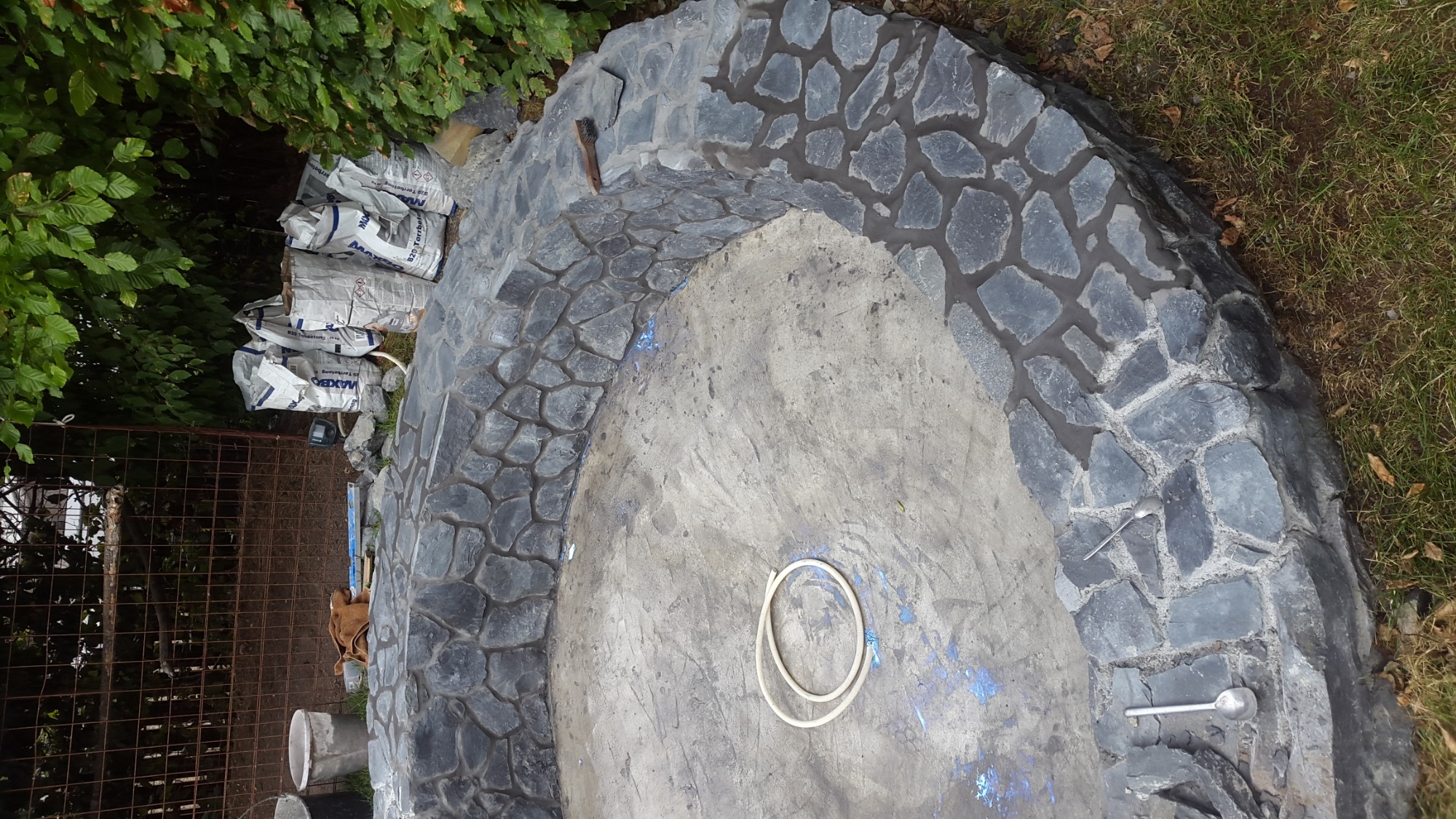 Budowa oczka wodnego: obrzeże z ciemnego kamienia, worki z zaprawą murarską w tle, widoczne narzędzia i wąż na betonowym dnie.