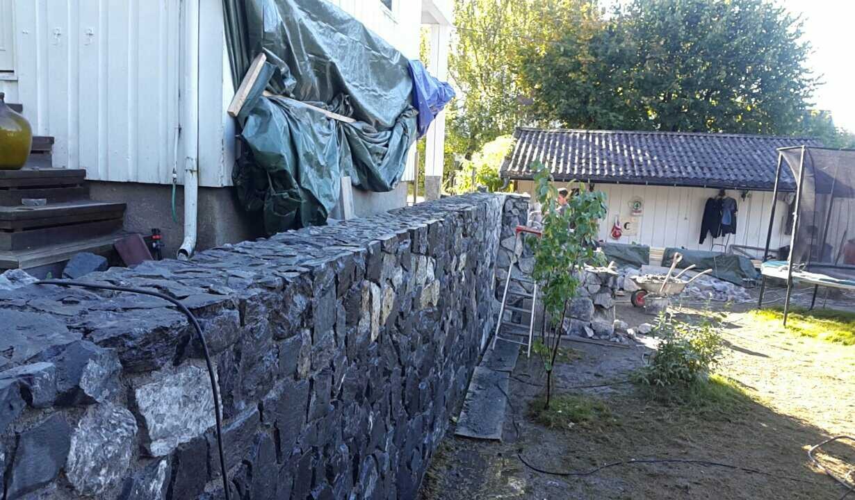 Wznoszenie kamiennego muru oporowego z nieregularnych, ciemnych kamieni, widoczne rusztowanie i narzędzia, częściowo zakryte plandeką, w tle biały budynek gospodarczy.