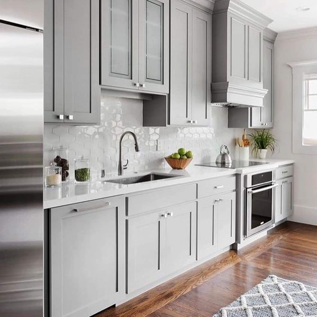 Jasnoszara kuchnia z białym heksagonalnym backsplashiem, drewnianą podłogą i dywanem w romby; owoce w drewnianej misce na blacie.