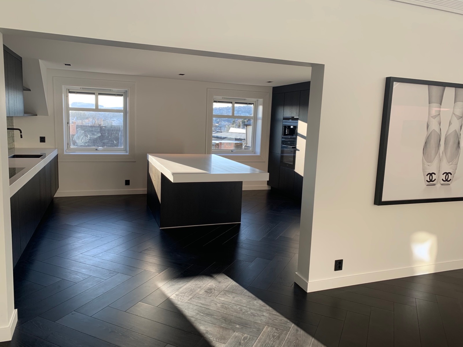 Nowoczesna kuchnia z ciemną, parkietową podłogą ułożoną w jodełkę, wyspą z białym blatem i czarną zabudową kuchenną, oświetlona promieniami słońca wpadającymi przez okno, z obrazem baletek Chanel...