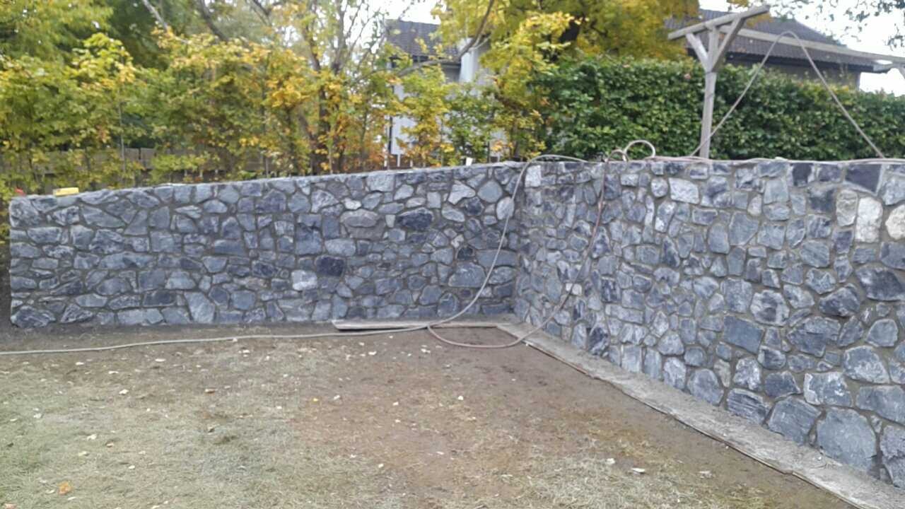 Kamienny mur ogrodowy z szarych kamieni różnej wielkości, widoczny kabel zasilający i drewniany element konstrukcyjny w tle.