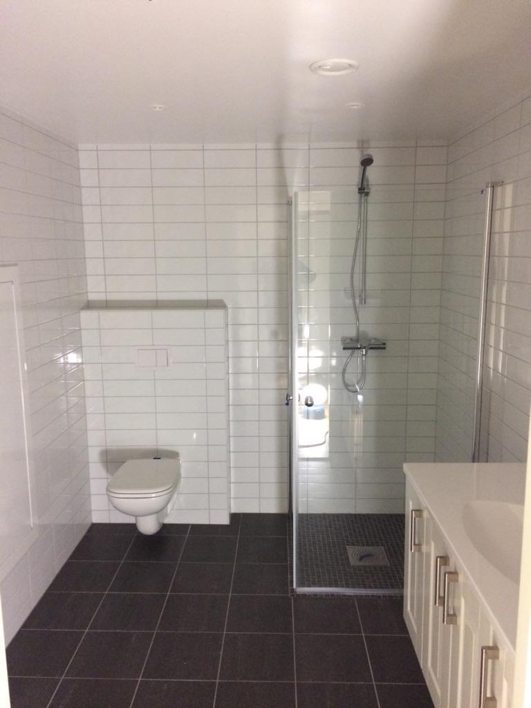 Nowoczesna łazienka z białymi płytkami, ciemną podłogą i kabiną prysznicową ze szklanymi drzwiami. Umywalka z szafką w stylu angielskim.