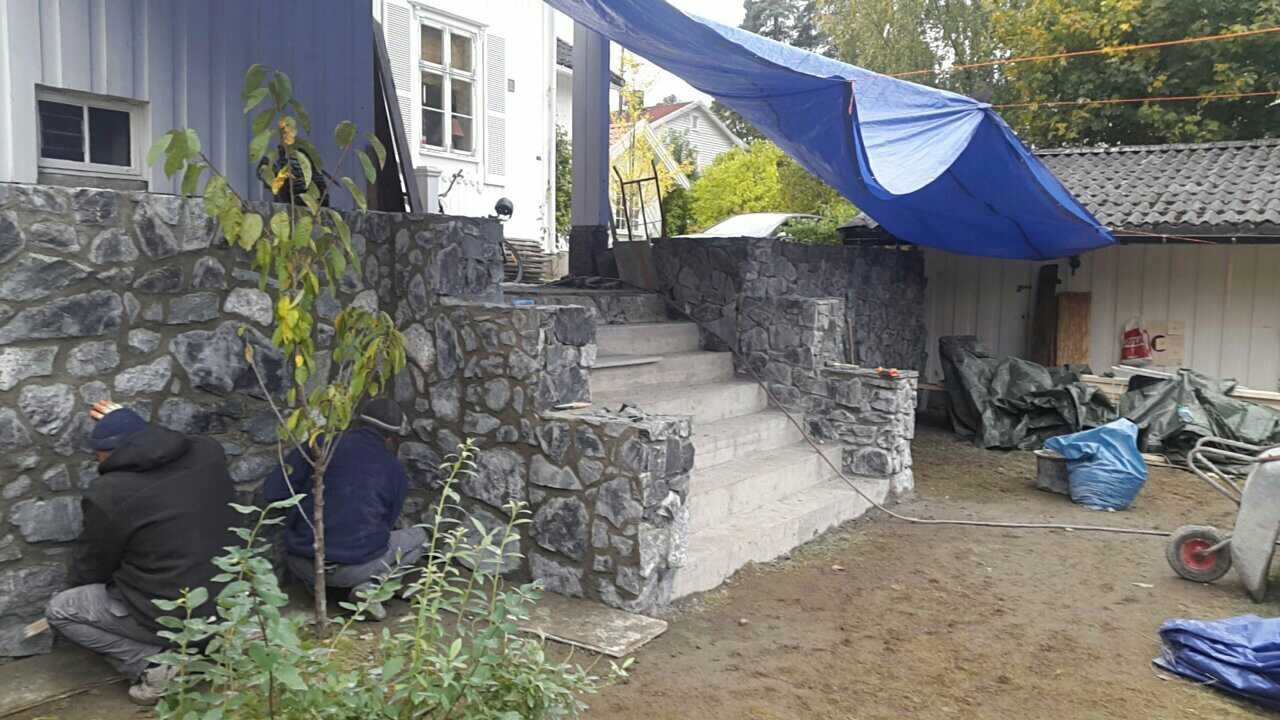 Kamienne schody wejściowe do domu w trakcie budowy, z widocznymi pracownikami i rozłożoną plandeką ochronną.