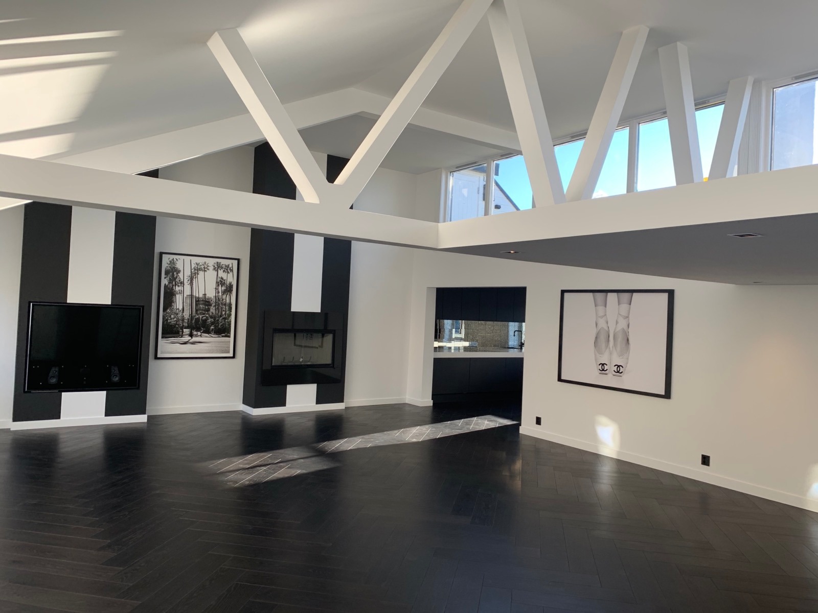 Przestronne wnętrze z ciemną podłogą ułożoną w jodełkę, białe ściany z czarnymi pasami, nowoczesny kominek, telewizor, grafiki w czarnych ramach i widok na kuchnię w tle.