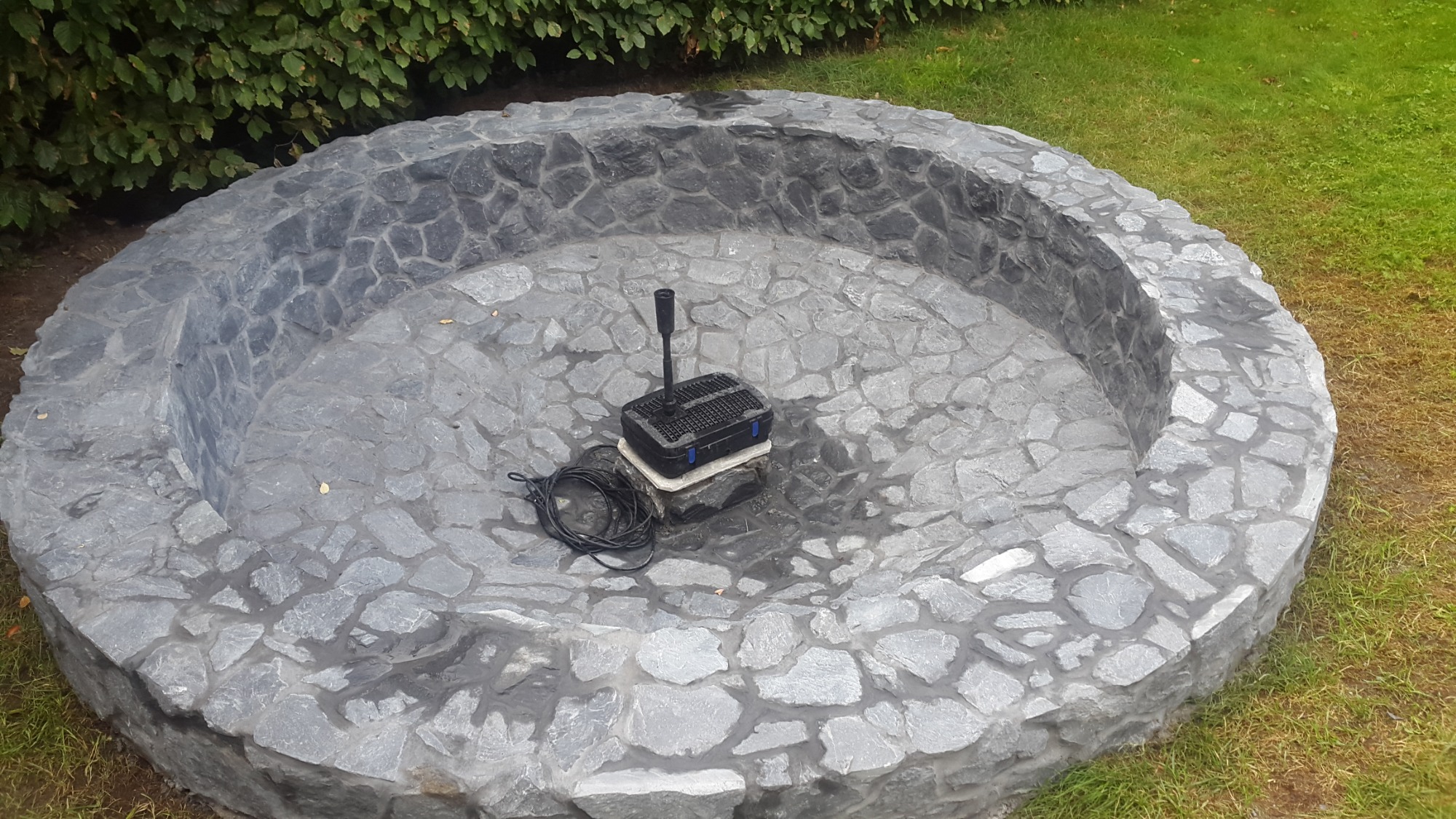 Okrągły, kamienny zbiornik w ogrodzie z zainstalowanym filtrem wodnym, otoczony trawnikiem i krzewami.