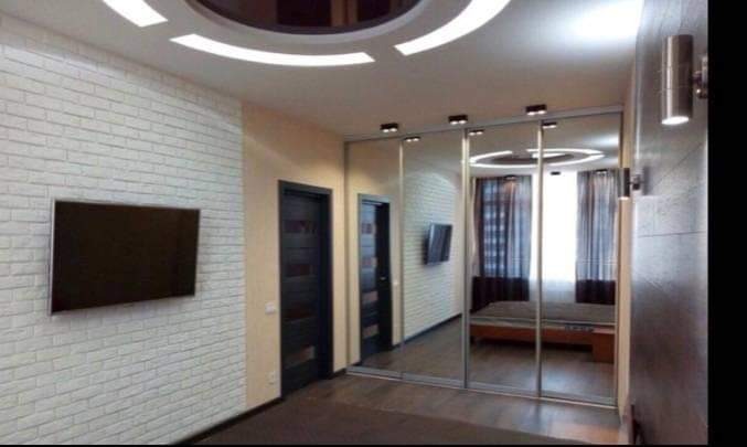 Nowoczesne wnętrze pokoju z białą ceglaną ścianą, telewizorem, przesuwną szafą z lustrami odbijającymi łóżko i okno, oraz designerskim oświetleniem sufitowym.