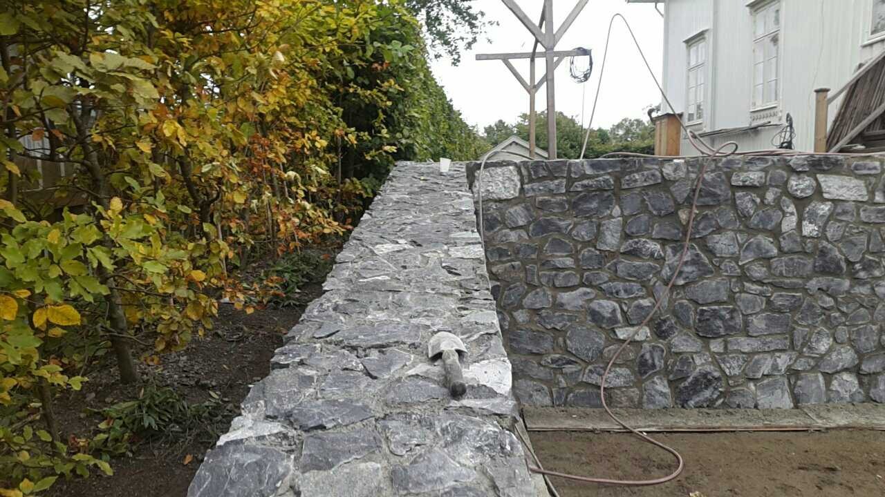 Kamienny murek w trakcie budowy, widoczny fragment budynku w tle, obok rosną drzewa z żółtymi liśćmi, na murku leży kielnia.