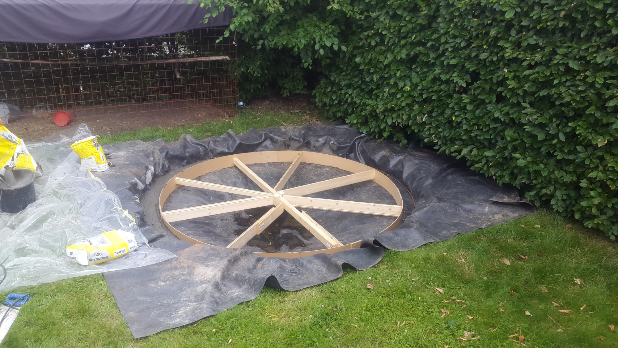 Przygotowanie do wylewki fundamentu pod okrągły element w ogrodzie, widoczna drewniana konstrukcja szalunku na czarnej folii, obok worki z zaprawą i narzędzia.