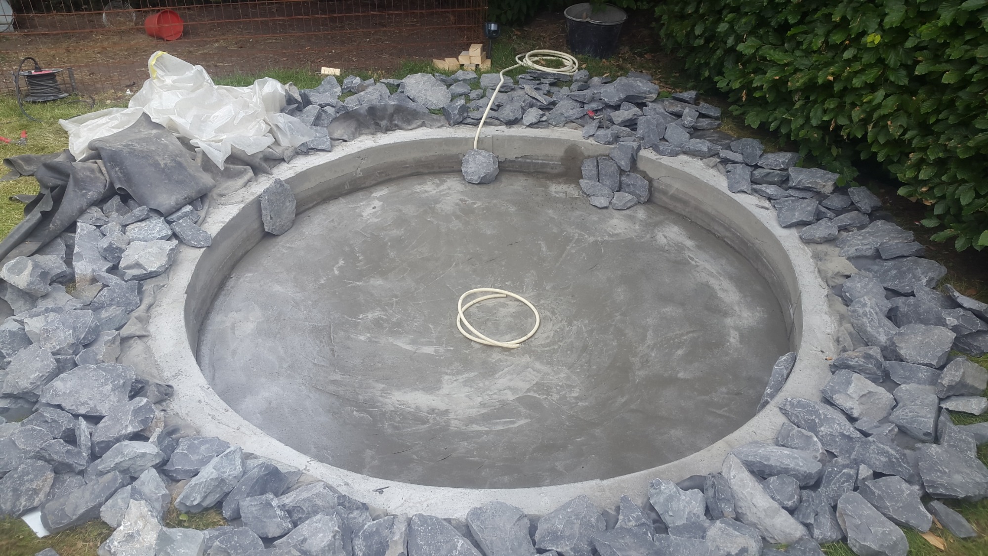 Betonowy okrągły zbiornik w trakcie budowy, obłożony szarymi kamieniami, widoczne narzędzia i materiały budowlane w tle.