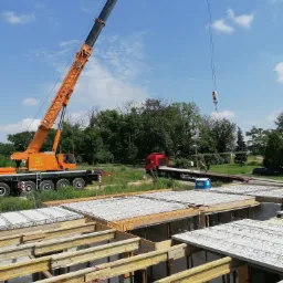 Żuraw budowlany podnosi belkę stalową nad fundamentami domu w trakcie budowy, widoczny transport ciężarowy na tle zieleni.
