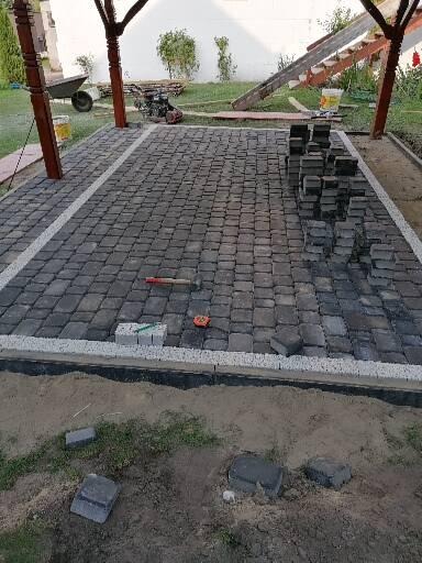 Układanie kostki brukowej na tarasie pod zadaszeniem, widoczne ułożone fragmenty, stosy kostki, narzędzia i przygotowane podłoże z piaskiem.