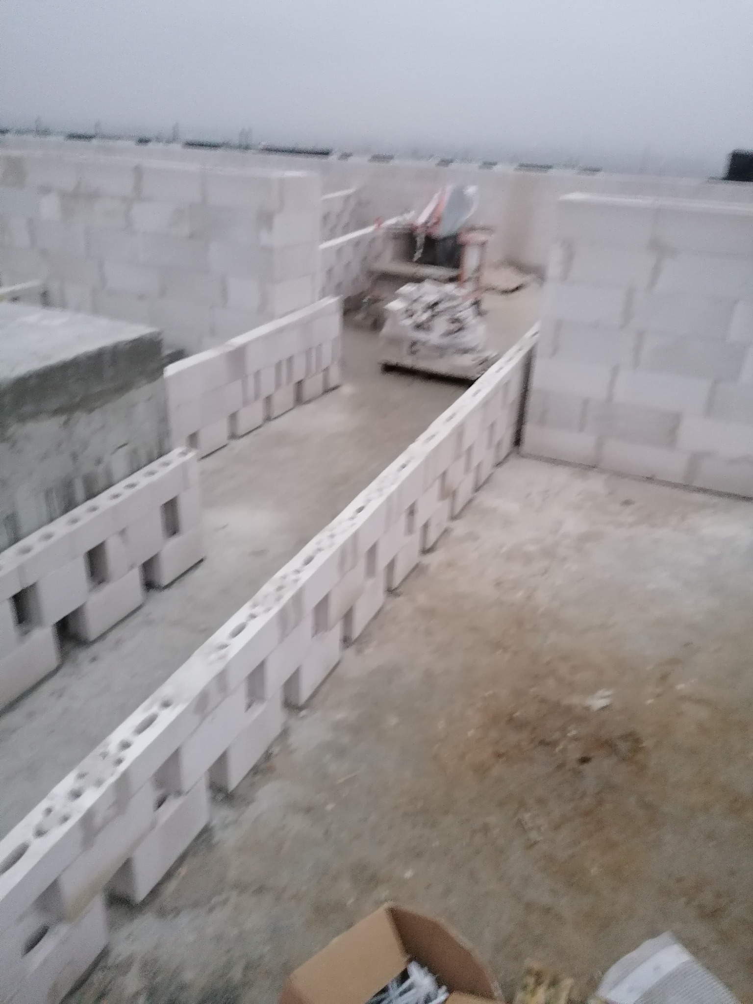 Wznoszenie ścian z białych bloczków betonu komórkowego Ytong na budowie domu, widoczne puste przestrzenie w bloczkach, w tle materiały budowlane pod folią.