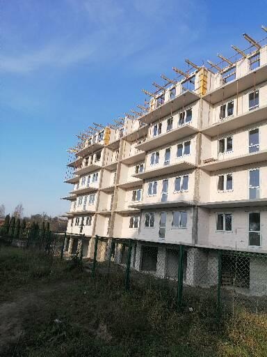 Wielopiętrowy budynek w stanie surowym otwartym, z widocznymi oknami i elementami konstrukcyjnymi na dachu, ogrodzony siatką.