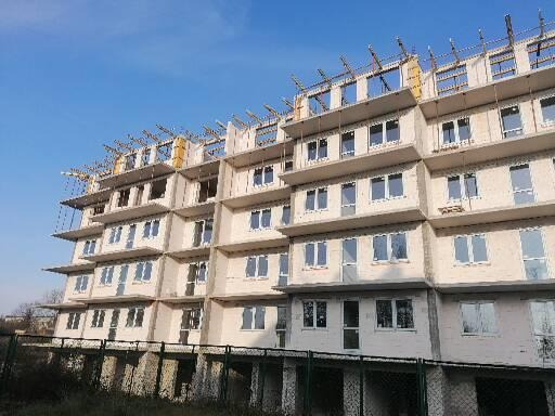 Wielopiętrowy budynek mieszkalny w trakcie budowy, widoczne surowe ściany, okna i drewniane szalunki na dachu, ogrodzenie siatkowe w dolnej części kadru.