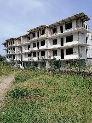 Wielopiętrowy budynek mieszkalny w stanie surowym, z widocznymi elementami konstrukcyjnymi, ogrodzony zieloną siatką, otoczony niezagospodarowanym terenem z bujną roślinnością.