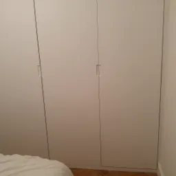 Biała szafa wnękowa z trzema drzwiami, widoczna z boku, obok białe drzwi wejściowe i fragment łóżka z białą pościelą.