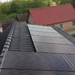 Panele słoneczne zamontowane na dachu z ciemnej dachówki ceramicznej, w tle budynki i drzewa.