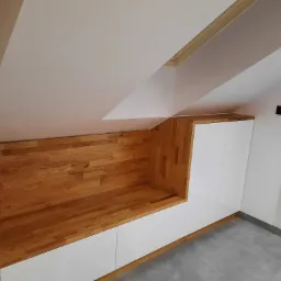 Drewniana zabudowa wnęki na poddaszu z białymi szafkami i półką, widoczny grzejnik drabinkowy na ścianie.