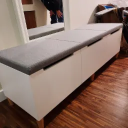 Biała szafka z szarym siedziskiem na drewnianych nóżkach, odbicie osoby w lustrze w tle, na parkiecie.