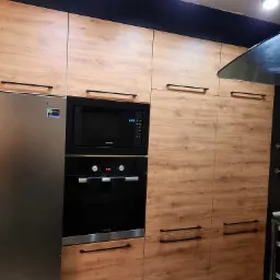 Nowoczesna zabudowa kuchenna z drewnopodobnymi frontami, czarnymi uchwytami, piekarnikiem i mikrofalówką marki Bosch i Samsung, oraz lodówką Samsung z widoczną naklejką gwarancyjną.