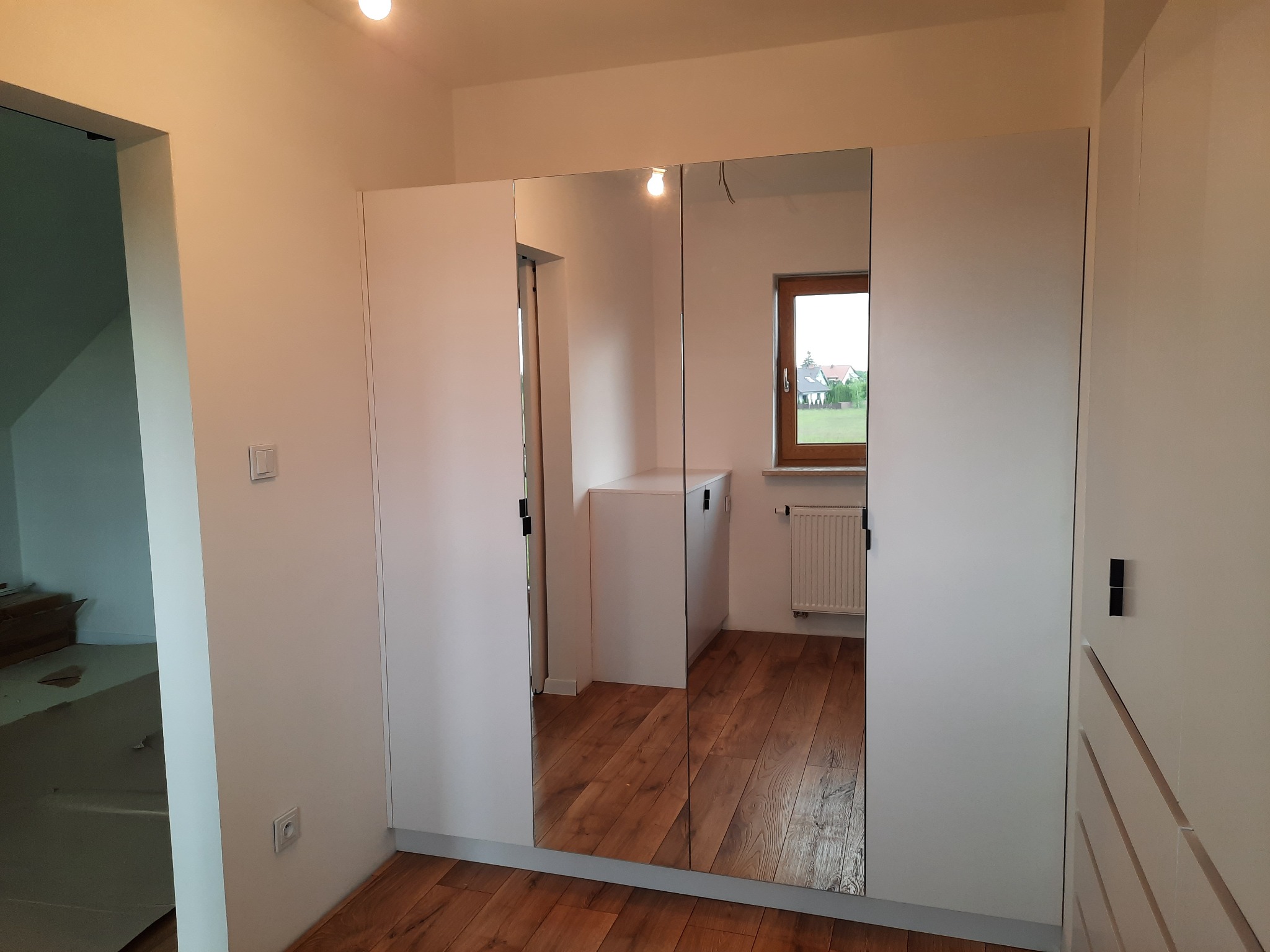 Biała szafa z lustrzanymi drzwiami w jasnym pomieszczeniu, widoczny parkiet na podłodze i okno z widokiem na domy w tle.