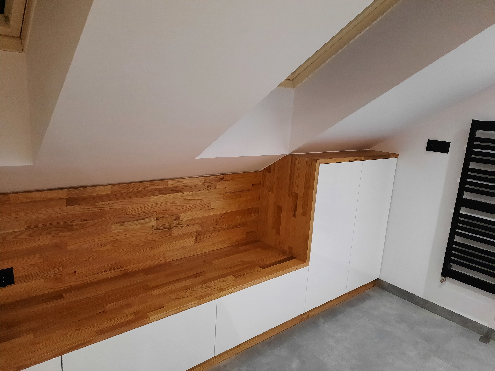 Drewniana zabudowa wnęki na poddaszu z białymi szafkami i półką, widoczny grzejnik drabinkowy na ścianie.