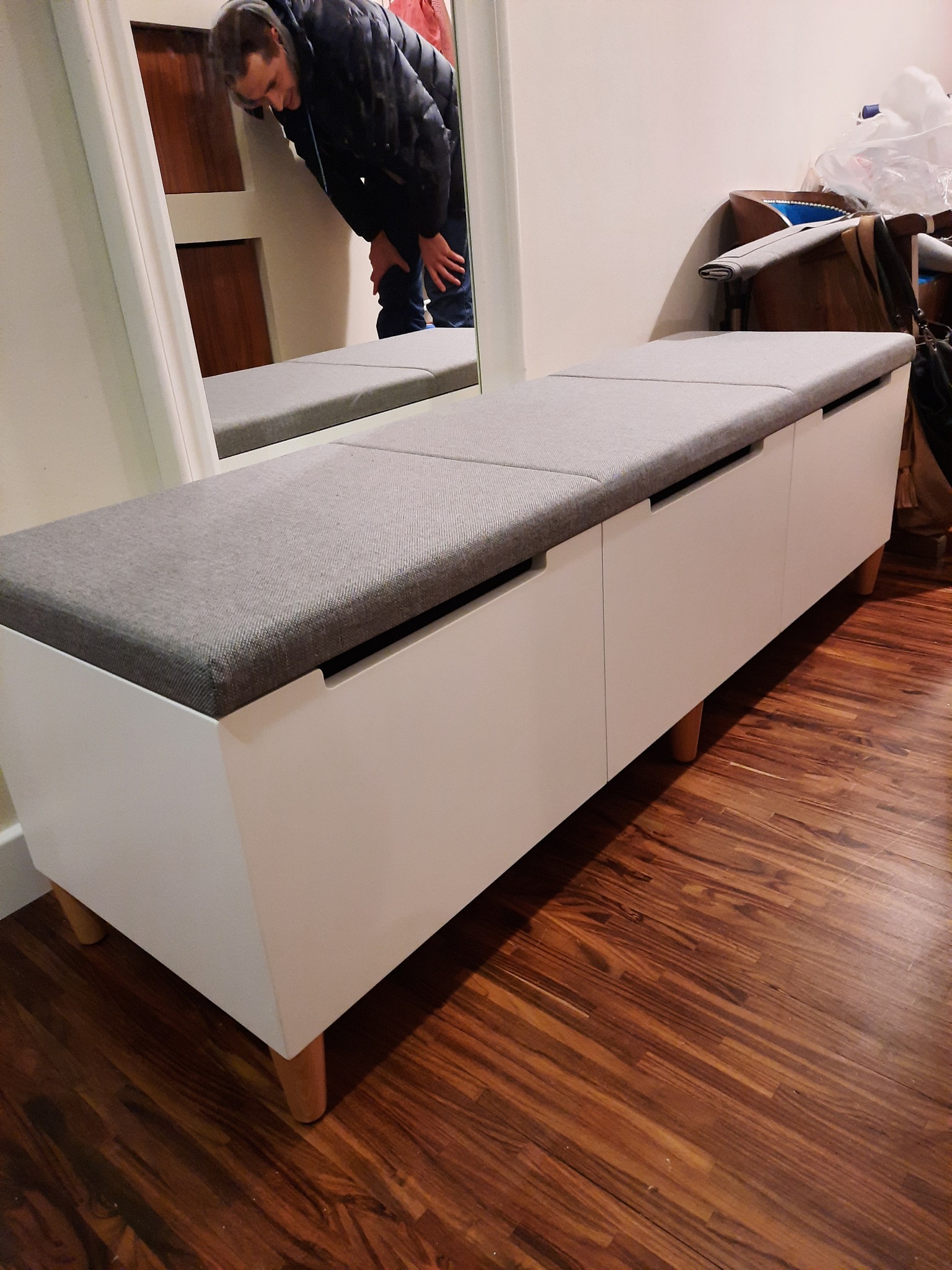 Biała szafka z szarym siedziskiem na drewnianych nóżkach, odbicie osoby w lustrze w tle, na parkiecie.