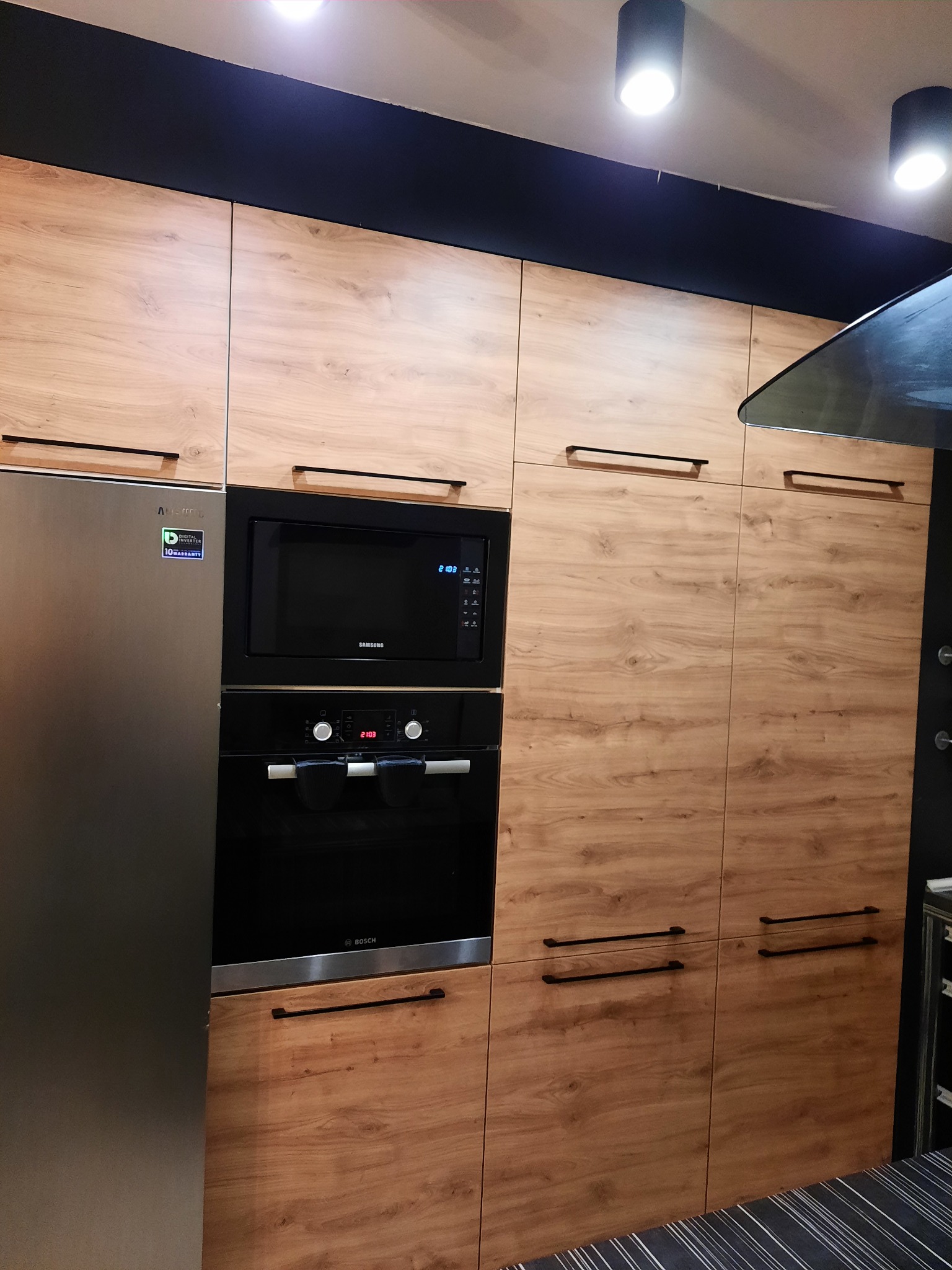 Nowoczesna zabudowa kuchenna z drewnopodobnymi frontami, czarnymi uchwytami, piekarnikiem i mikrofalówką marki Bosch i Samsung, oraz lodówką Samsung z widoczną naklejką gwarancyjną.