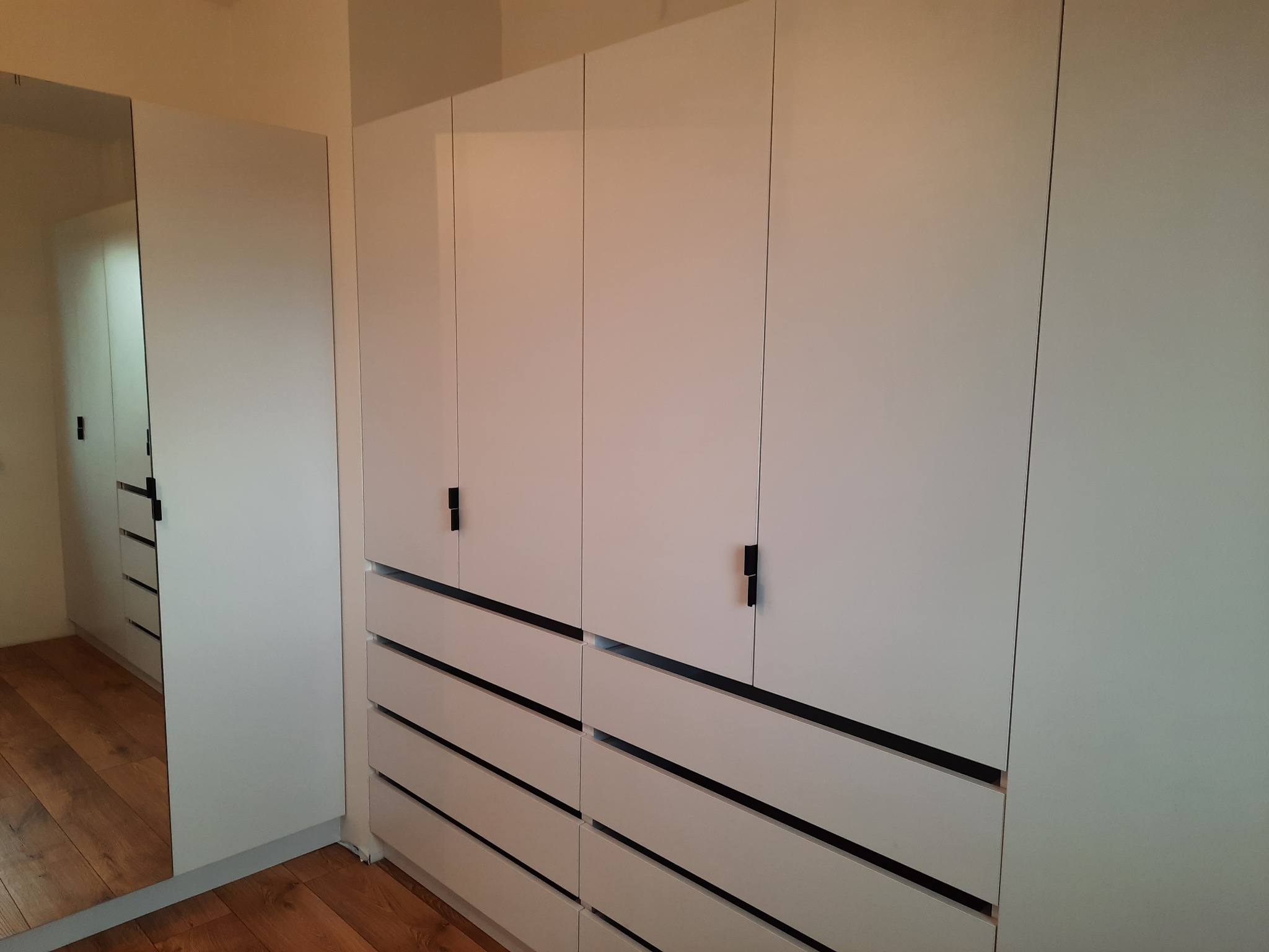 Biała szafa wnękowa z czarnymi uchwytami i szufladami, widoczne odbicie parkietu w lustrzanych drzwiach.