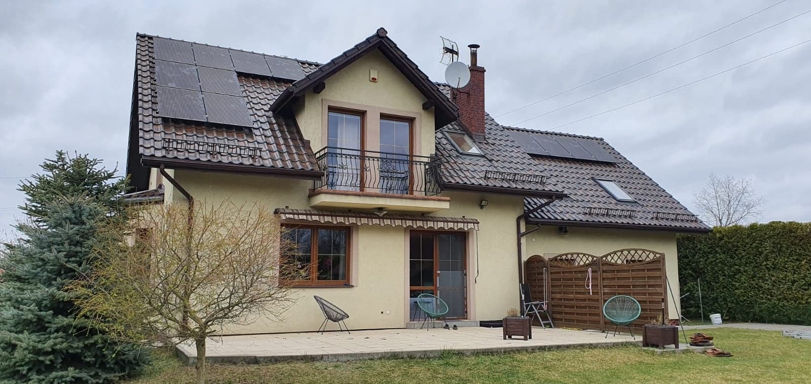 Dom jednorodzinny z panelami słonecznymi na dachu, widocznymi z przodu i z tyłu, z anteną satelitarną i kominem z czerwonej cegły, na tle pochmurnego nieba.